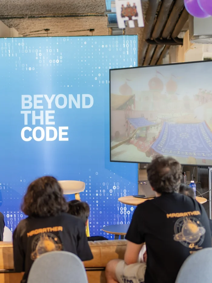 Audience seated at a tech event in front of a blue backdrop that reads ‘BEYOND THE CODE’ with a large display screen to the right and balloons overhead. Audience seated and watching a presentation in a room; a large blue banner reads 'BEYOND THE CODE' on the left.