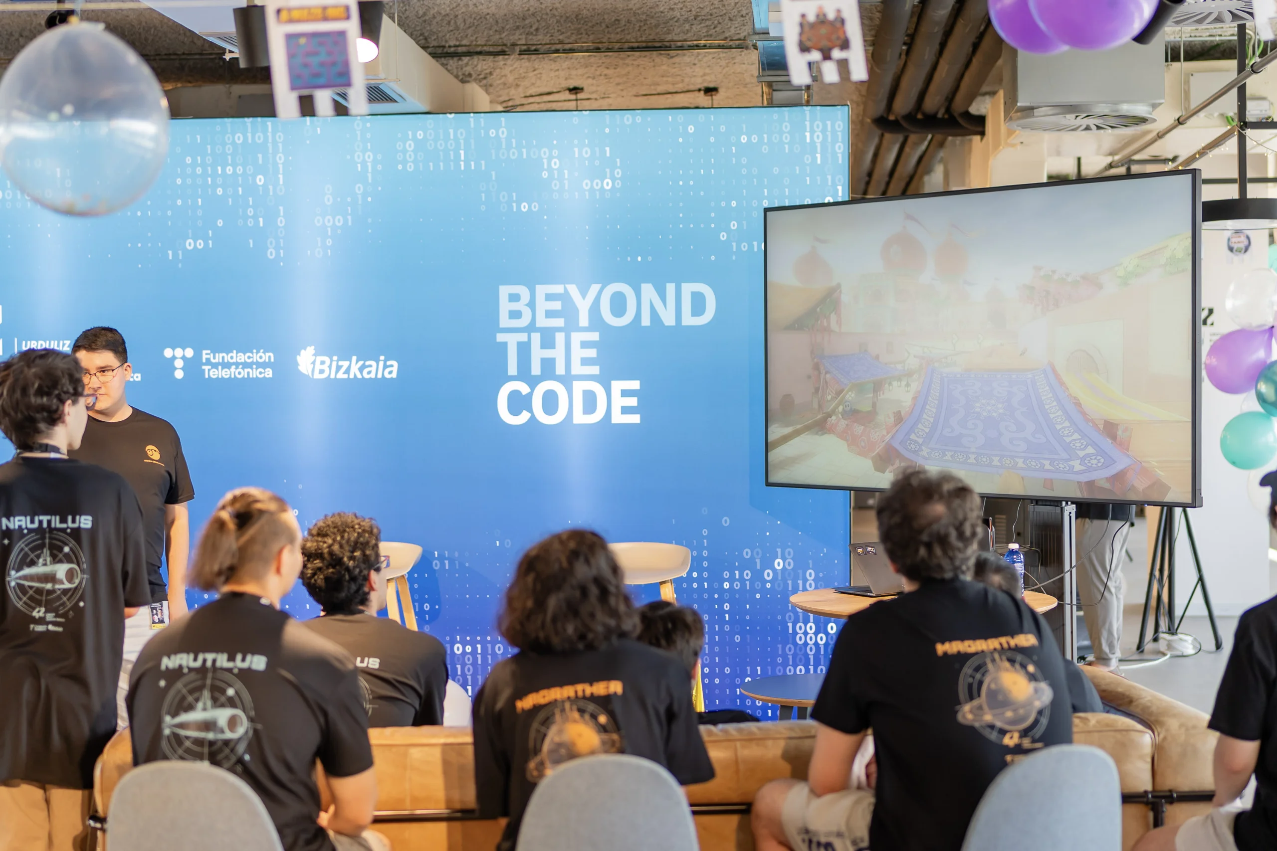 Audience seated at a tech event in front of a blue backdrop that reads 'BEYOND THE CODE' with a large display screen to the right and balloons overhead.